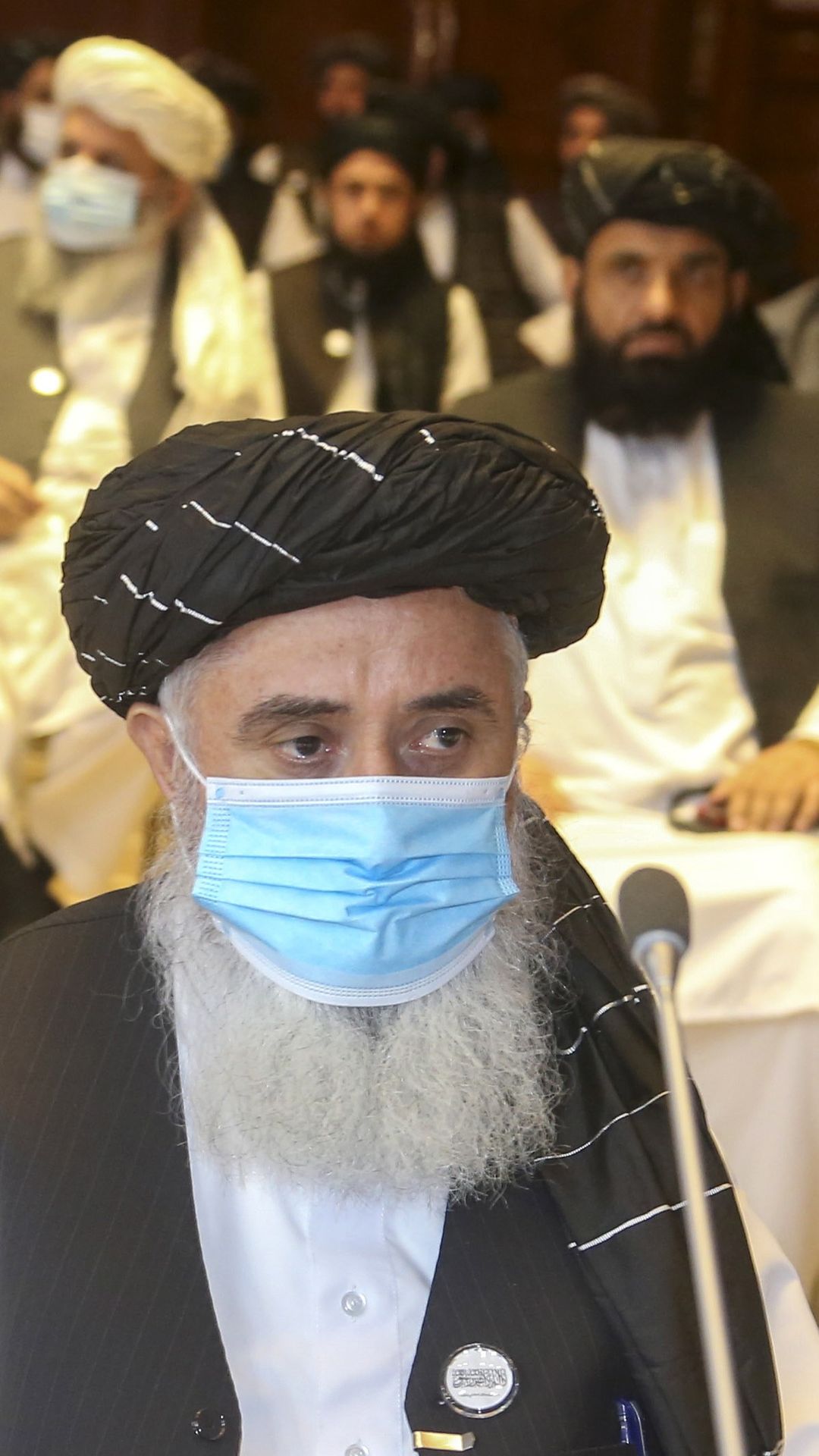 Talibanin edustaja, jolla on pyöreä päähine, harmaa parta ja kertakäyttömaski, istuu salissa Dohassa. Taustalla istuu valkoisiin pukeutuneita, parrakkaita miehiä.