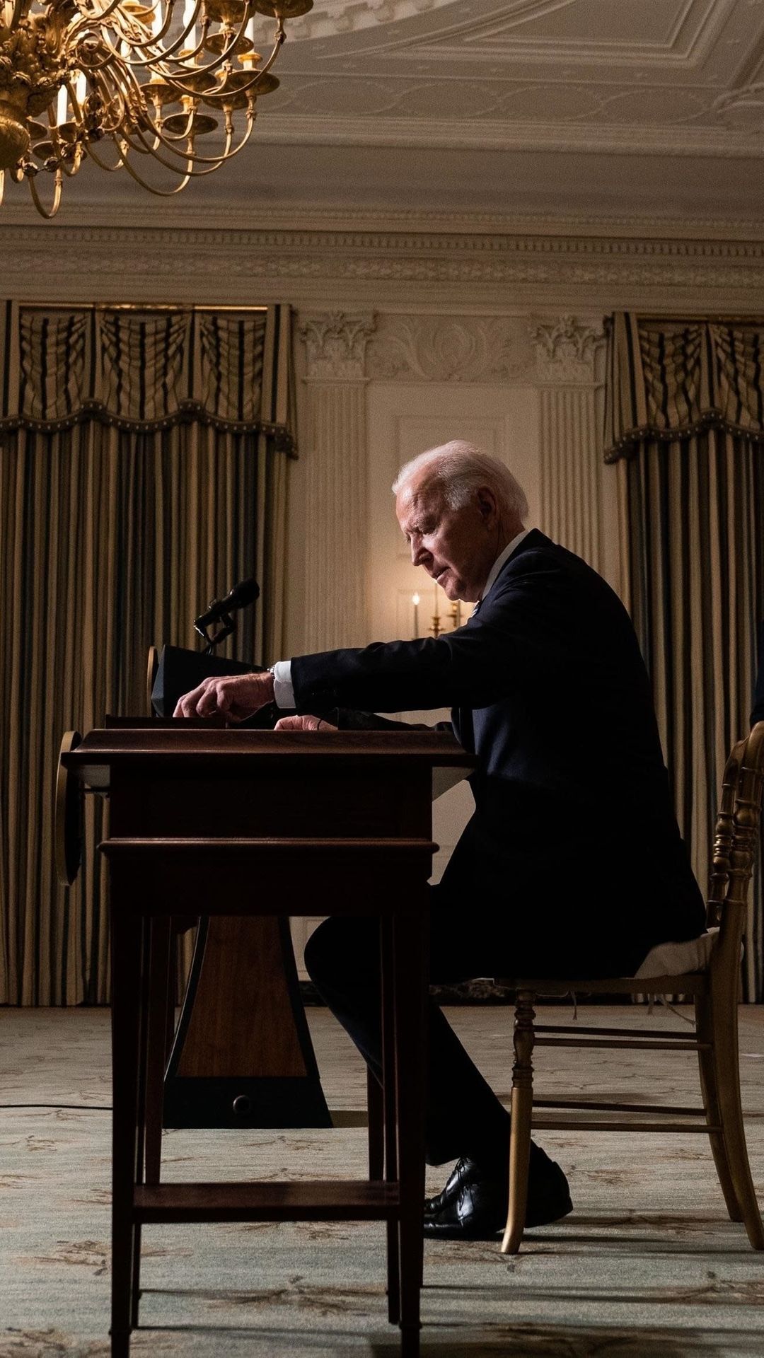 Joe Biden kuvattuna sivusta, kun hän istuu työpöydän ääressä ja allekirjoittaa papereita suuressa huoneessa..