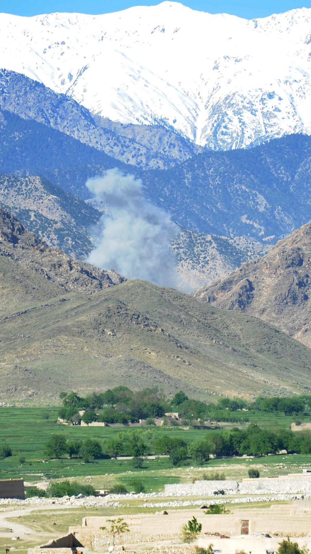 Savupilvi nousee keskeltä vuoria, joiden korkeimmat huiput ovat lumisia. Etualalla näkyy kaistale vihreää metsikköä ja talojen kattoja.