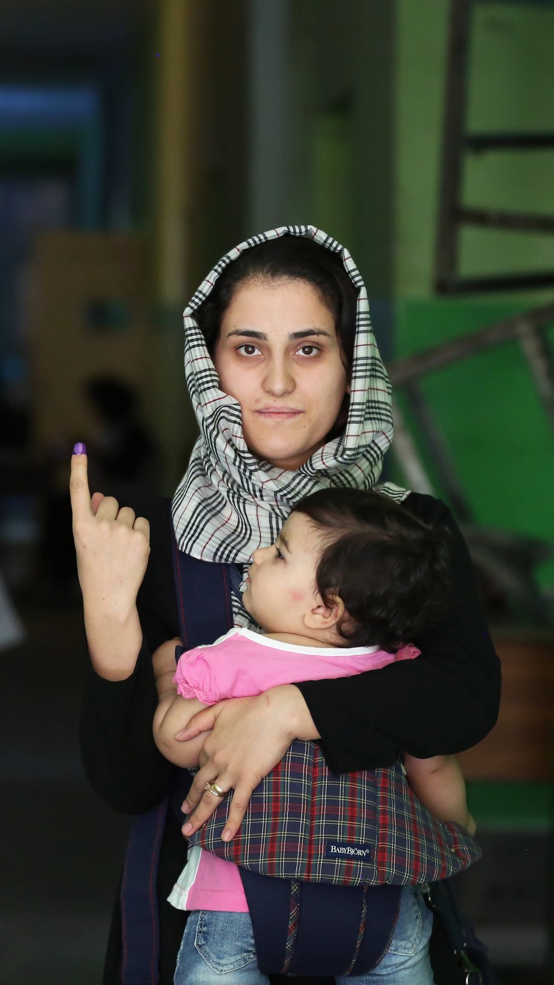 Nainen näyttää etusormeaan, joka on kastettu violettiin maaliin äänestämisen jälkeen. Hänellä on sylissään mustatukkainen, pinkkipuseroinen pieni lapsi. 