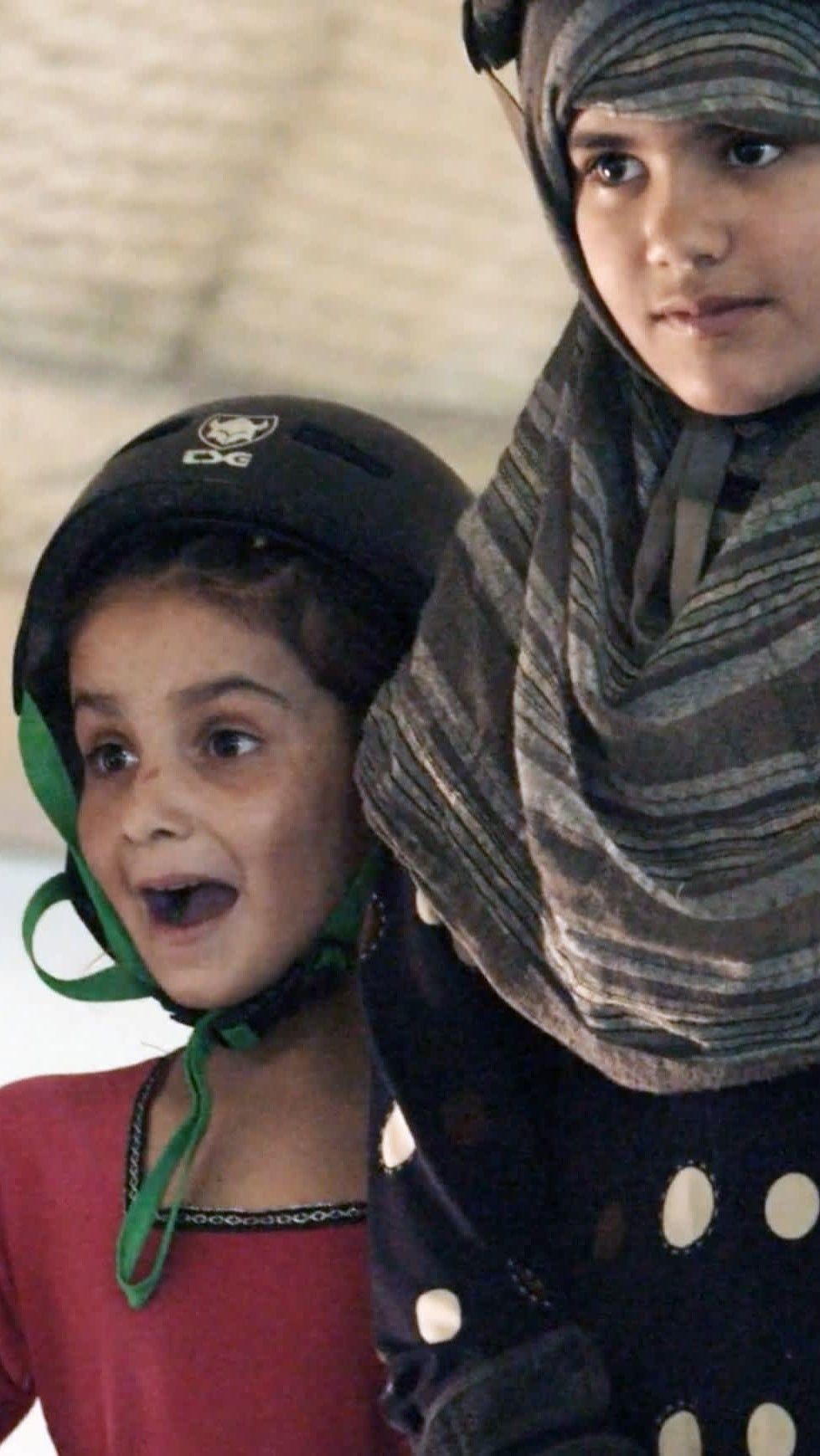 Kaksi kypäräpäistä skeittaavaa tyttöä, joista toisella on huivi kypärän alla ja toisella innostunut ilme. 