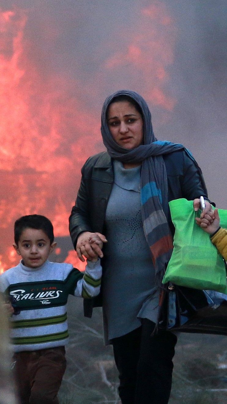 Huivipäiinen nainen pitää kädestä kiinni mustatukkaista pientä poikaa, taustalla on tulimeri. 