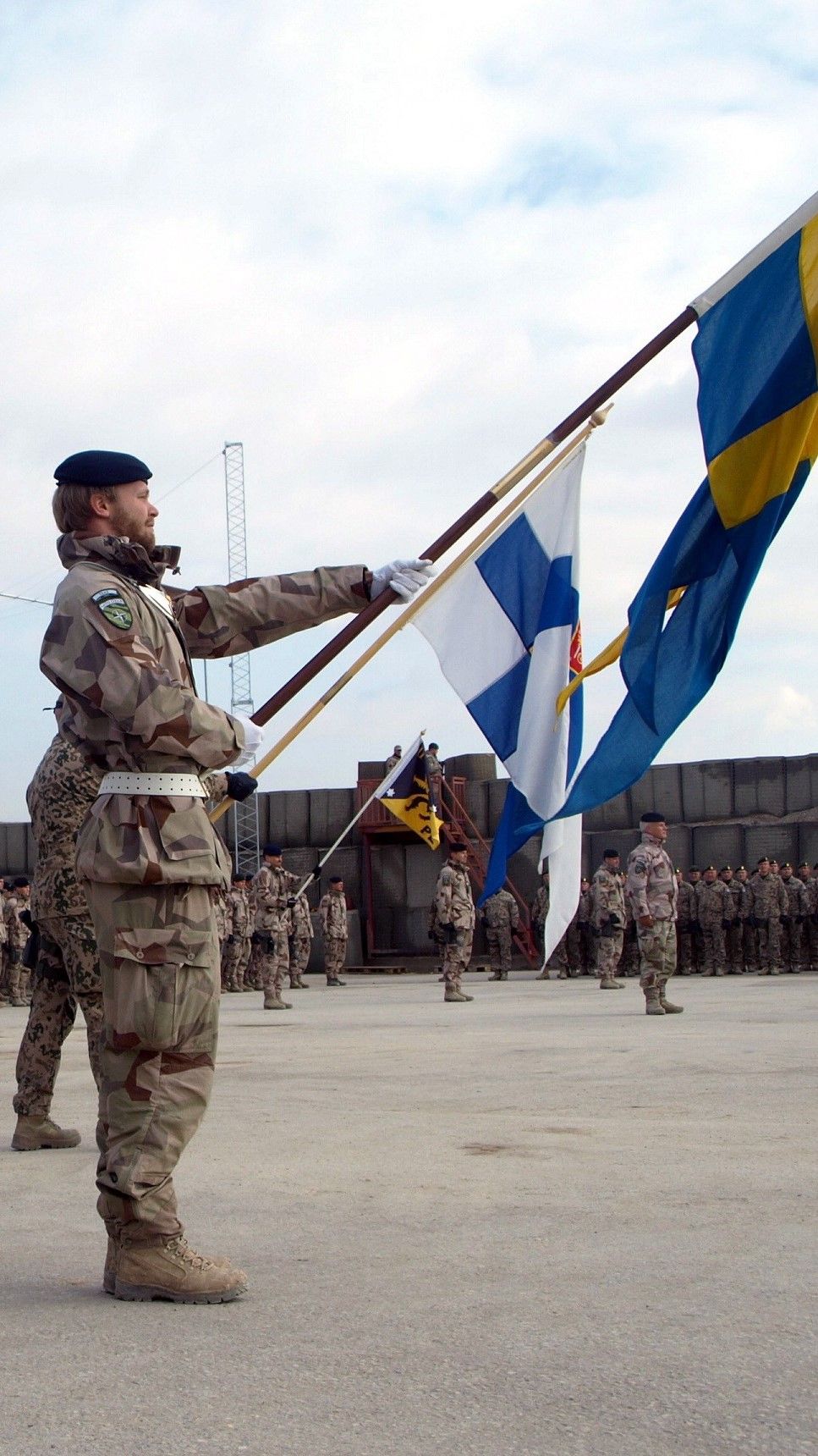 Ruotsalainen ja suomalainen sotilas pitävät maiden lippuja tanassa. Taustalla seisoo muita sotilasasuisia henkilöitä. 