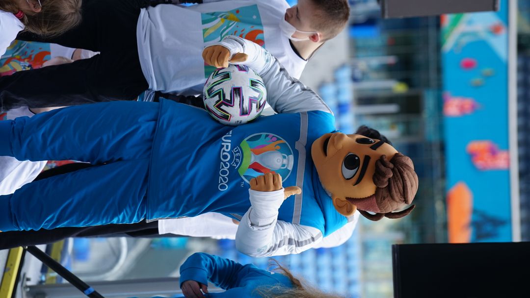 Nuorta miestä esittävä kisamaskotti näyttää peukkua.